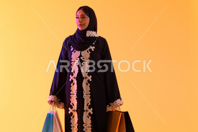 بورترية لسيده عربية خليجية اماراتيه مبتسمة ترتدي العباءه والحجاب تحمل بيدها أكياس التسوق الملونه، الأثر الإيجابي للتسوق على السيدات، سيدة سعودية تستفيد من العروض الموسمية والتخفيضات،  التبضع والتسوق من الماركات العالميه، خلفية صفراء