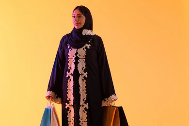 A portrait of a smiling Emirati Gulf Arab woman wearing an abaya and hijab, holding colorful shopping bags in her hand, the positive impact of shopping on women, a Saudi woman benefiting from seasonal offers and discounts, shopping from global brands, yellow background.