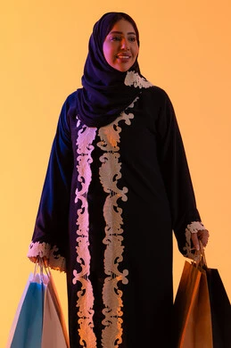 A Saudi woman benefits from seasonal offers and discounts, shopping from global brands, a portrait of a smiling Arab Gulf Emirati woman wearing an abaya and hijab holding colorful shopping bags in her hand, the positive impact of shopping on women, yellow background.
