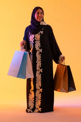 الأثر الإيجابي للتسوق على السيدات، سيدة سعودية تستفيد من العروض الموسمية والتخفيضات،  التبضع والتسوق من الماركات العالميه، بورترية لسيده عربية خليجية اماراتيه مبتسمة ترتدي العباءه والحجاب تحمل بيدها أكياس التسوق الملونه، ، خلفية صفراء