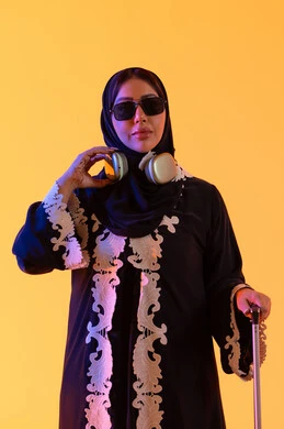 سماع الموسيقي عبر الهيدفون ،ارتداء النظارات الشمسية ، الاستعداد و تجهيز الأمتعة للسفر في جولة سياحية، مفهوم السياحة و السفر، بورتريه لامراه سعوديه مبتسمه تمسك حقيبة سفر ، امراه عربيه خليجيه اماراتيه ترتدي العباءه المطرزة والحجاب تنظر بايماءات وجه تدل على الحماس للقيام برحلة سياحية،  خلفية صفراء