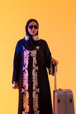 الاستعداد و تجهيز الأمتعة للسفر في جولة سياحية، مفهوم السياحة و السفر، سماع الموسيقي عبر الهيدفون ،ارتداء النظارات الشمسية ،بورتريه لامراه سعوديه مبتسمه تمسك حقيبة سفر ، امراه عربيه خليجيه اماراتيه ترتدي العباءه المطرزة والحجاب تنظر بايماءات وجه تدل على الحماس للقيام برحلة سياحية،  خلفية صفراء