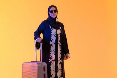ارتداء النظارات الشمسية ، الاستعداد و تجهيز الأمتعة للسفر في جولة سياحية، مفهوم السياحة و السفر، بورتريه لامراه سعوديه مبتسمه تمسك حقيبة سفر ، امراه عربيه خليجيه اماراتيه ترتدي العباءه المطرزة والحجاب تنظر بايماءات وجه تدل على الحماس للقيام برحلة سياحية،  خلفية صفراء
