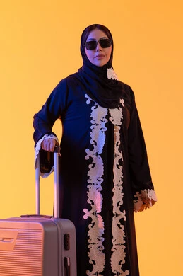 امراه عربيه خليجيه اماراتيه ترتدي العباءه المطرزة والحجاب تنظر بايماءات وجه تدل على الحماس للقيام برحلة سياحية، ارتداء النظارات الشمسية ، الاستعداد و تجهيز الأمتعة للسفر في جولة سياحية، مفهوم السياحة و السفر، بورتريه لامراه سعوديه مبتسمه تمسك حقيبة سفر ،  خلفية صفراء