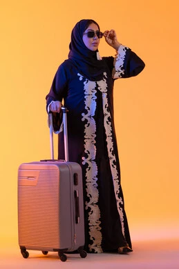 بورتريه لامراه سعوديه مبتسمه تمسك حقيبة سفر ، امراه عربيه خليجيه اماراتيه ترتدي العباءه المطرزة والحجاب تنظر بايماءات وجه تدل على الحماس للقيام برحلة سياحية، ارتداء النظارات الشمسية ، الاستعداد و تجهيز الأمتعة للسفر في جولة سياحية، مفهوم السياحة و السفر،  خلفية صفراء