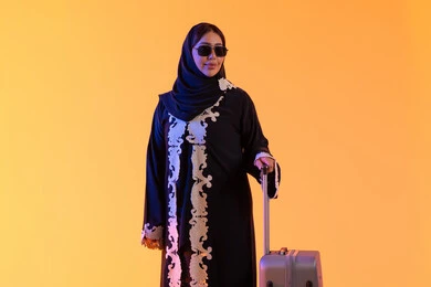 مفهوم السياحة و السفر، بورتريه لامراه سعوديه مبتسمه تمسك حقيبة سفر ، ارتداء النظارات الشمسية ، الاستعداد و تجهيز الأمتعة للسفر في جولة سياحية، امراه عربيه خليجيه اماراتيه ترتدي العباءه المطرزة والحجاب تنظر بايماءات وجه تدل على الحماس للقيام برحلة سياحية،  خلفية صفراء