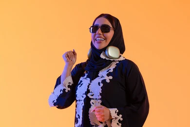 The concept of listening and enjoyment, an Emirati woman using headphones in her free time, a portrait of a Saudi Gulf Arab woman wearing an embroidered abaya and hijab, putting on headphones with expressions of happiness, the development of technology and tech, spending enjoyable time listening to music, yellow background.