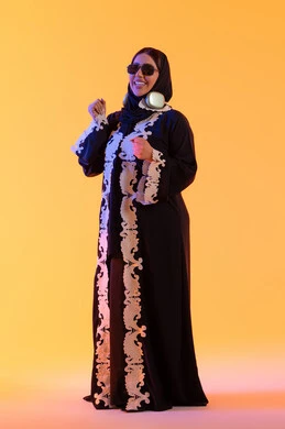 قضاء وقت ممتع بالاستماع الى الموسيقى, مفهوم الإصغاء والاستمتاع، سيدة اماراتية تستخدم الهيدفون في اوقات الفراغ، بورتريه لامرأة عربية خليجية سعودية ترتدي العباءة المطرزة والحجاب تضع سماعات الرأس بتعابير السعادة، تطور التقنيه والتكنولوجيا، خلفية صفراء