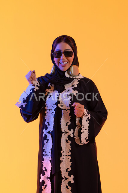 رفع قبضة اليد بايماءات الحماس ،قضاء وقت ممتع بالاستماع الى الموسيقى, مفهوم الإصغاء والاستمتاع، سيدة اماراتية تستخدم الهيدفون في اوقات الفراغ، بورتريه لامرأة عربية خليجية سعودية ترتدي العباءة المطرزة والحجاب تضع سماعات الرأس بتعابير السعادة، تطور التقنيه والتكنولوجيا، خلفية صفراء