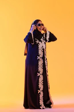 تطور التقنيه والتكنولوجيا، قضاء وقت ممتع بالاستماع الى الموسيقى, مفهوم الإصغاء والاستمتاع، سيدة اماراتية تستخدم الهيدفون في اوقات الفراغ، بورتريه لامرأة عربية خليجية سعودية ترتدي العباءة المطرزة والحجاب تضع سماعات الرأس بتعابير السعادة، خلفية صفراء