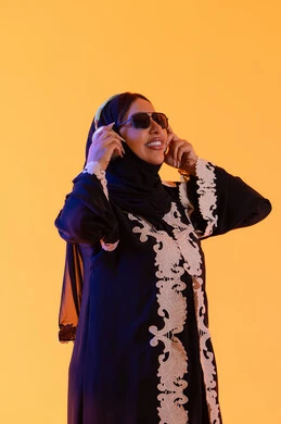 بورتريه لامرأة عربية خليجية سعودية ترتدي العباءة المطرزة والحجاب تضع سماعات الرأس بتعابير السعادة، تطور التقنيه والتكنولوجيا، قضاء وقت ممتع بالاستماع الى الموسيقى, مفهوم الإصغاء والاستمتاع، سيدة اماراتية تستخدم الهيدفون في اوقات الفراغ، خلفية صفراء