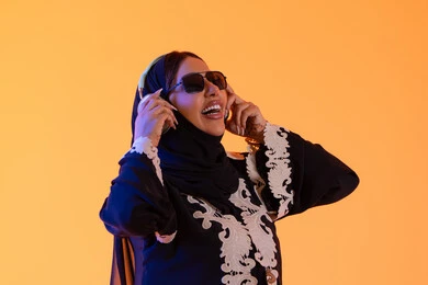 An Emirati woman using headphones in her free time, a portrait of a Gulf Arab Saudi woman wearing an embroidered abaya and hijab, putting on headphones with expressions of happiness, the development of technology and tech, enjoying time listening to music, the concept of listening and enjoyment, yellow background.