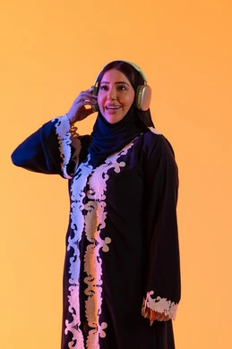 تطور التقنيه والتكنولوجيا، قضاء وقت ممتع بالاستماع الى الموسيقى, مفهوم الإصغاء والاستمتاع، سيدة اماراتية تستخدم الهيدفون في اوقات الفراغ، بورتريه لامرأة عربية خليجية سعودية ترتدي العباءة المطرزة والحجاب تضع سماعات الرأس بتعابير السعادة، خلفية صفراء