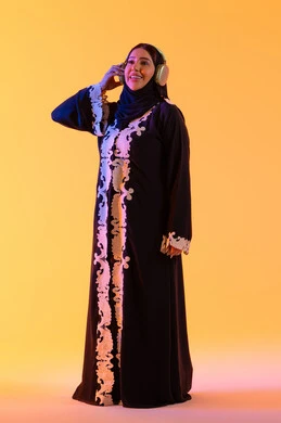 بورتريه لامرأة عربية خليجية سعودية ترتدي العباءة المطرزة والحجاب تضع سماعات الرأس بتعابير السعادة، تطور التقنيه والتكنولوجيا،قضاء وقت ممتع بالاستماع الى الموسيقى, مفهوم الإصغاء والاستمتاع، سيدة اماراتية تستخدم الهيدفون في اوقات الفراغ،  خلفية صفراء