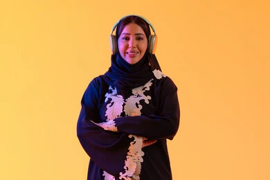 سيدة اماراتية تستخدم الهيدفون في اوقات الفراغ، بورتريه لامرأة عربية خليجية سعودية ترتدي العباءة المطرزة والحجاب تضع سماعات الرأس بتعابير السعادة، تطور التقنيه والتكنولوجيا،قضاء وقت ممتع بالاستماع الى الموسيقى, مفهوم الإصغاء والاستمتاع،  خلفية صفراء