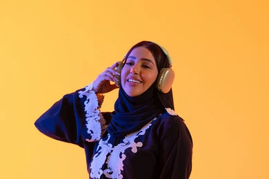 مفهوم الإصغاء والاستمتاع، سيدة اماراتية تستخدم الهيدفون في اوقات الفراغ، بورتريه لامرأة عربية خليجية سعودية ترتدي العباءة المطرزة والحجاب تضع سماعات الرأس بتعابير السعادة، تطور التقنيه والتكنولوجيا، قضاء وقت ممتع بالاستماع الى الموسيقى, خلفية صفراء
