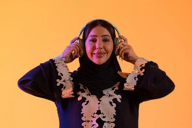 بورتريه لامرأة عربية خليجية سعودية ترتدي العباءة المطرزة والحجاب تضع سماعات الرأس بتعابير السعادة، تطور التقنيه والتكنولوجيا،قضاء وقت ممتع بالاستماع الى الموسيقى, مفهوم الإصغاء والاستمتاع، سيدة اماراتية تستخدم الهيدفون في اوقات الفراغ،  خلفية صفراء