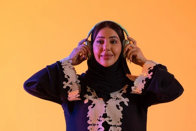 سيدة اماراتية تستخدم الهيدفون في اوقات الفراغ، بورتريه لامرأة عربية خليجية سعودية ترتدي العباءة المطرزة والحجاب تضع سماعات الرأس بتعابير السعادة، تطور التقنيه والتكنولوجيا،قضاء وقت ممتع بالاستماع الى الموسيقى, مفهوم الإصغاء والاستمتاع،  خلفية صفراء