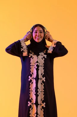 مفهوم الإصغاء والاستمتاع، سيدة اماراتية تستخدم الهيدفون في اوقات الفراغ، بورتريه لامرأة عربية خليجية سعودية ترتدي العباءة المطرزة والحجاب تضع سماعات الرأس بتعابير السعادة، تطور التقنيه والتكنولوجيا، قضاء وقت ممتع بالاستماع الى الموسيقى, خلفية صفراء
