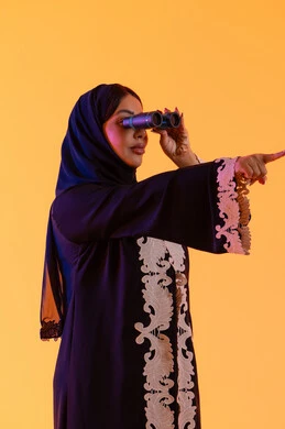 رؤية الاشياء البعيدة وتقريبها، الاشاره الى شئ ما ، سيده سعوديه تحب الاستكشاف والمغامرة، بورتريه لامراه عربيه خليجيه اماراتيه ترتدي العباءه والحجاب تحمل بيدها منظار ثنائي العينين وتنظر بإيماءات السرور والاستمتاع ، خلفية صفراء