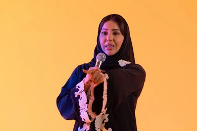 بورتريه لامراه عربيه خليجيه اماراتيه ترتدي العباءه المطرزة والحجاب تمسك بيدها المايكروفون ، سيده سعوديه تغني بإيماءات الحماس والسرور، تعابير الاندماج والتفاعل مع الموسيقي، تقديم العروض الصوتية ، تسجيل مقطع صوتي غنائي، تقديم البرامج الاذاعية، ممارسة موهبة الغناء،خلفية صفراء