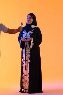 تقديم البرامج الاذاعية،العمل في تقديم الفقرات و البرامج الفنية الترفيهية، بورتريه لامراه عربيه خليجيه اماراتيه ترتدي العباءه المطرزة والحجاب تمسك بيدها المايكروفون ، سيده سعوديه تقدم اخبار عالميه متنوعه بإيماءات السرور، تعابير الاندماج والتفاعل ، تقديم العروض الصوتية ، خلفية صفراء