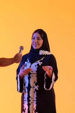سيده سعوديه تقدم اخبار عالميه متنوعه بإيماءات السرور، تعابير الاندماج والتفاعل ، العمل في تقديم الفقرات و البرامج الفنية الترفيهية، تقديم البرامج الاذاعية، بورتريه لامراه عربيه خليجيه اماراتيه ترتدي العباءه المطرزة والحجاب تمسك بيدها المايكروفون ، تقديم العروض الصوتية ، خلفية صفراء