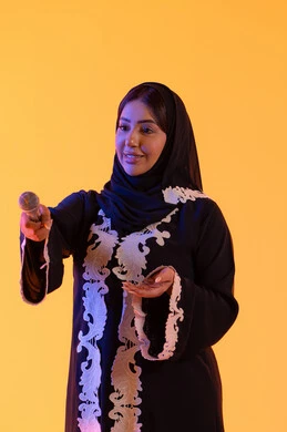 تقديم العروض الصوتية ، تسجيل مقطع صوتي غنائي، تقديم البرامج الاذاعية، ممارسة موهبة الغناء، بورتريه لامراه عربيه خليجيه اماراتيه ترتدي العباءه المطرزة والحجاب تمسك بيدها المايكروفون ، سيده سعوديه تغني بإيماءات الحماس والسرور، تعابير الاندماج والتفاعل مع الموسيقي، خلفية صفراء