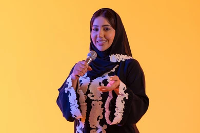تقديم البرامج الاذاعية، تسجيل مقطع صوتي غنائي، ممارسة موهبة الغناء، بورتريه لامراه عربيه خليجيه اماراتيه ترتدي العباءه المطرزة والحجاب تمسك بيدها المايكروفون ، سيده سعوديه تغني بإيماءات الحماس والسرور، تعابير الاندماج والتفاعل مع الموسيقي، تقديم العروض الصوتية ، خلفية صفراء
