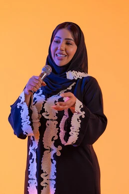 تسجيل مقطع صوتي غنائي، تقديم البرامج الاذاعية، ممارسة موهبة الغناء، بورتريه لامراه عربيه خليجيه اماراتيه ترتدي العباءه المطرزة والحجاب تمسك بيدها المايكروفون ، سيده سعوديه تغني بإيماءات الحماس والسرور، تعابير الاندماج والتفاعل مع الموسيقي، تقديم العروض الصوتية ، خلفية صفراء
