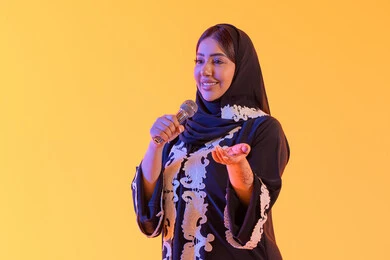سيده سعوديه تغني بإيماءات الحماس والسرور، تعابير الاندماج والتفاعل مع الموسيقي، تقديم العروض الصوتية ، تسجيل مقطع صوتي غنائي، تقديم البرامج الاذاعية، ممارسة موهبة الغناء، بورتريه لامراه عربيه خليجيه اماراتيه ترتدي العباءه المطرزة والحجاب تمسك بيدها المايكروفون ، خلفية صفراء