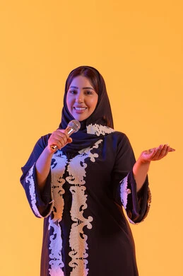 تقديم العروض الصوتية ، تسجيل مقطع صوتي غنائي، تقديم البرامج الاذاعية، ممارسة موهبة الغناء، بورتريه لامراه عربيه خليجيه اماراتيه ترتدي العباءه المطرزة والحجاب تمسك بيدها المايكروفون ، سيده سعوديه تغني بإيماءات الحماس والسرور، تعابير الاندماج والتفاعل مع الموسيقي، خلفية صفراء
