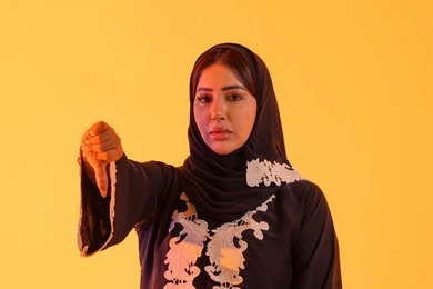 بورتريه لامراه عربيه خليجيه اماراتيه ترتدي العباءه والحجاب تقفف بايماءات يد تدل على عدم الموافقه وعدم الإعجاب والرفض ، سيدة سعودية تشير بأصبع الإبهام لأسفل, الإنزعاج من شيء ما ،خلفية صفراء