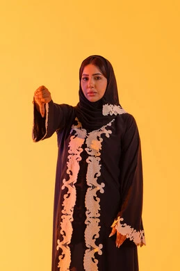A Saudi woman pointing her thumb down, showing discomfort about something, a portrait of an Arab Gulf Emirati woman wearing an abaya and hijab, standing with hand gestures indicating disagreement, disapproval, and rejection, with a yellow background.