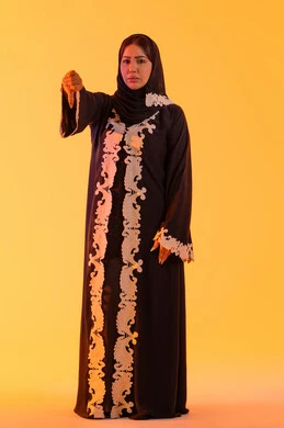 سيدة سعودية تشير بأصبع الإبهام لأسفل, الإنزعاج من شيء ما ، بورتريه لامراه عربيه خليجيه اماراتيه ترتدي العباءه والحجاب تقفف بايماءات يد تدل على عدم الموافقه وعدم الإعجاب والرفض ، خلفية صفراء