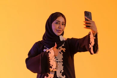 الدردشة والتحدث مع الأصدقاء، اجراء مكالمه فيديو ، تصفح مواقع وتطبيقات الانترنت، بورتريه لامراه عربيه خليجيه اماراتيه ترتدي العباءه والحجاب تمسك الجوال بيدها، سيده سعوديه تستخدم التقنيات والتكنولوجيا الحديثة بالحياة اليومية، خلفية صفراء