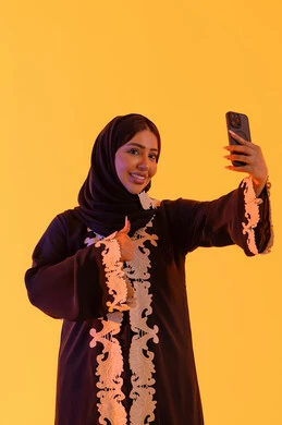 Making a video call, chatting and talking with friends, browsing websites and internet applications, a portrait of an Arab Gulf Emirati woman wearing an abaya and hijab holding a mobile phone in her hand, a Saudi woman using modern technologies and technology in daily life, yellow background.