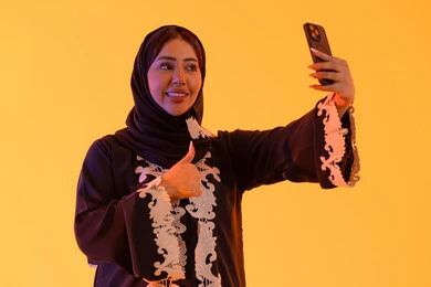 ايماءات الجودة والرضي ، اجراء مكالمه فيديو ، الدردشة والتحدث مع الأصدقاء،تصفح مواقع وتطبيقات الانترنت، بورتريه لامراه عربيه خليجيه اماراتيه ترتدي العباءه والحجاب تمسك الجوال بيدها، سيده سعوديه تستخدم التقنيات والتكنولوجيا الحديثة بالحياة اليومية، خلفية صفراء