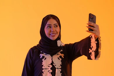 بورتريه لامراه عربيه خليجيه اماراتيه ترتدي العباءه والحجاب تمسك الجوال بيدها، اجراء مكالمه فيديو ، الدردشة والتحدث مع الأصدقاء،تصفح مواقع وتطبيقات الانترنت،سيده سعوديه تستخدم التقنيات والتكنولوجيا الحديثة بالحياة اليومية، خلفية صفراء