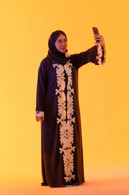 Browsing websites and internet applications, making video calls, chatting and talking with friends, a portrait of an Arab Gulf Emirati woman wearing an abaya and hijab holding a mobile phone in her hand, a Saudi woman using modern technologies and technology in daily life, yellow background.
