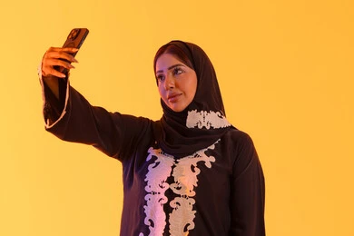 Chatting and talking with friends, making a video call, browsing websites and internet applications, a portrait of an Arab Gulf Emirati woman wearing an abaya and hijab holding a mobile phone in her hand, a Saudi woman using modern technologies and technology in daily life, yellow background.