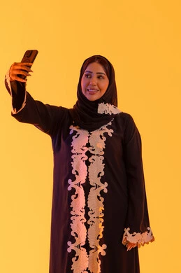 بورتريه لامراه عربيه خليجيه اماراتيه ترتدي العباءه والحجاب تمسك الجوال بيدها، اجراء مكالمه فيديو ، الدردشة والتحدث مع الأصدقاء،تصفح مواقع وتطبيقات الانترنت،سيده سعوديه تستخدم التقنيات والتكنولوجيا الحديثة بالحياة اليومية، خلفية صفراء