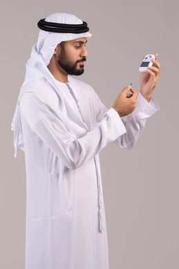 الإصابة بداء السكري، مشاكل ارتفاع السكر في الدم، بورتريه لشاب عربي خليجي اماراتي مريض يرتدي الكندورة والغترة الاماراتية يقوم بفحص نسبة السكر في دمه، أجهزة الكترونية حديثة للفحص الطبي السريع، خلفية بيضاء