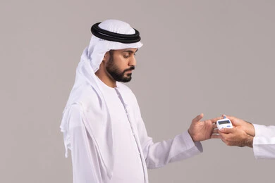 الإصابة بداء السكري، مشاكل ارتفاع السكر في الدم، بورتريه لشاب عربي خليجي اماراتي مريض يرتدي الكندورة والغترة الاماراتية يقوم بفحص نسبة السكر في دمه، أجهزة الكترونية حديثة للفحص الطبي السريع، خلفية بيضاء