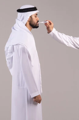 The need for medical assistance, the importance of having a digital scale at home, the feeling of fatigue and exhaustion and symptoms of fever, a side portrait of an Emirati Gulf Arab man wearing a kandura and ghutrah holding a thermometer measuring his temperature, white background.