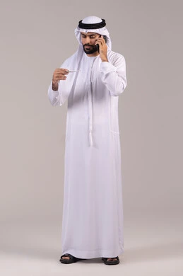 Making a phone call via mobile to consult the doctor about the appropriate treatment, a portrait of an Emirati Gulf Arab man wearing a kandura and ghutrah holding a thermometer and looking at the indicator, the need for medical assistance, feeling fatigued and symptoms of fever, full-body image, white background.