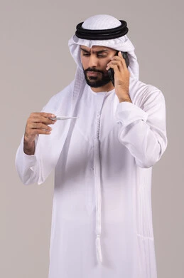 Feeling exhausted and having fever symptoms, making a phone call via mobile to consult a doctor about the appropriate treatment, a portrait of an Emirati Gulf Arab man wearing a kandura and ghutrah holding a thermometer and looking at the indicator, the need for medical assistance, white background. Feeling exhausted and having fever symptoms, making a phone call via mobile to consult a doctor about the appropriate treatment, a portrait of an Emirati Gulf Arab man wearing a kandura and ghutrah holding a thermometer and looking at the indicator, the need for medical assistance, white background.