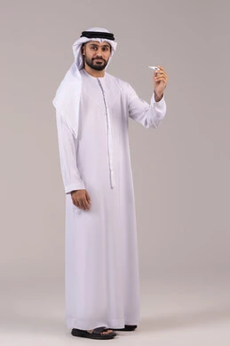 الشعور بالتحسن وانخفاض الحرارة, مفهوم التماثل للشفاء، بورتريه لرجل عربي خليجي اماراتي يرتدي الكندورة والغترة يمسك بيده جهاز مقياس الحرارة، النظر الى الكاميرا بتعابير السرور والفرح للتعافي من المرض، صورة الجسم بالطول الكامل, خلفية بيضاء