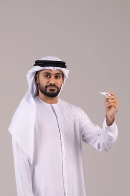 مفهوم التماثل للشفاء، الشعور بالتحسن وانخفاض الحرارة, بورتريه لرجل عربي خليجي اماراتي يرتدي الكندورة والغترة يمسك بيده جهاز مقياس الحرارة، النظر الى الكاميرا بتعابير السرور والفرح للتعافي من المرض، خلفية بيضاء