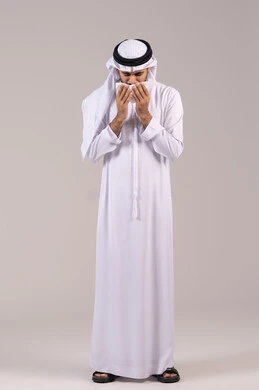 Nasal and sinus allergy inflammation, the need for treatment and medication, feeling unwell and having a cold, a portrait of an Emirati Gulf Arab man wearing a kandura and ghutrah holding a tissue to his face with sneezing gestures, full-body, white background.